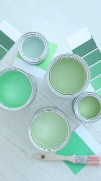 Metal cans with paint in shades of green, brush and color palettes on white wooden table, flat lay. Camera spinning