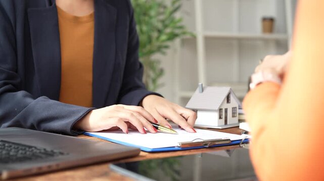 Real estate agent discussing a house sale contract with a client, explaining details and gesturing while reviewing the document in a professional office environment
