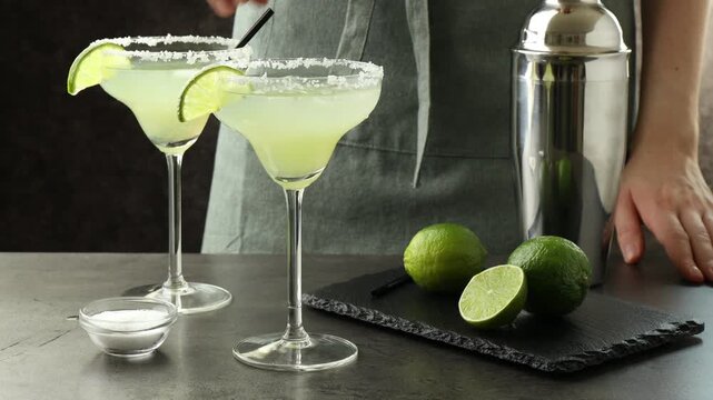 Woman putting straws in glasses of margarita cocktail and taking shaker at table, closeup