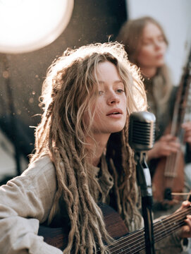 boho female musician with dreadlocks singing softly into vintage microphone while playing acoustic guitar, warm lens flare effect, ideal for indie music, acoustic concert, artistic performance