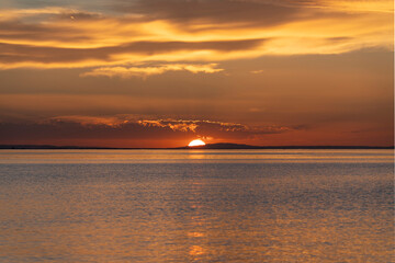 Fototapeta premium sunset over Lake Constance captured from the shoreline of Bregenz, Austria, with the setting sun dipping behind distant hills and casting a brilliant golden glow over the calm water