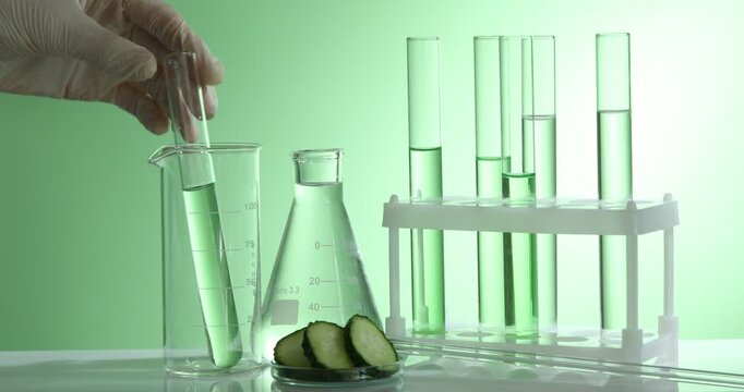 Woman putting test tubes with liquids into rack, pouring water in beaker and cucumber slices at table against green gradient background, closeup