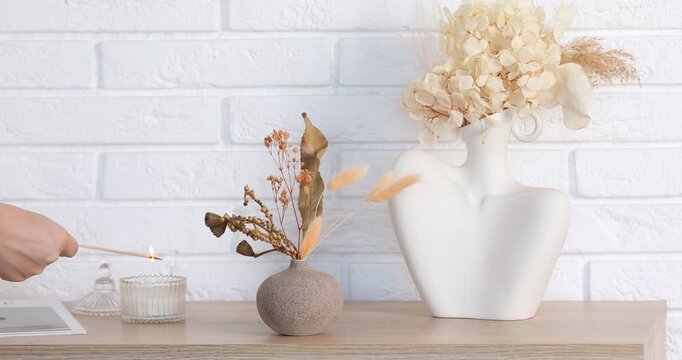 Woman lighting candle on wooden dresser near vases of dry flowers indoors, closeup