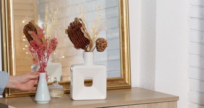 Woman putting beautiful dry flowers in vase onto wooden dresser indoors, closeup