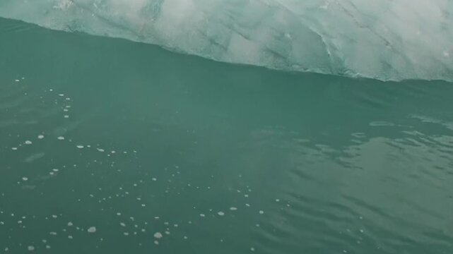 Blue ice and irregular frozen texture fill a close natural view. The frame also shows surface reflections, calm conditions and green water, adding visible texture and spatial context to the scene.