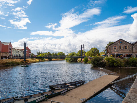 Cricklepit Suspension Bridge crosses the River Exe at the historic Exeter Quay, a popular waterside leisure location in Devon. Canoes are moored tat a pontoon in the foreground. 