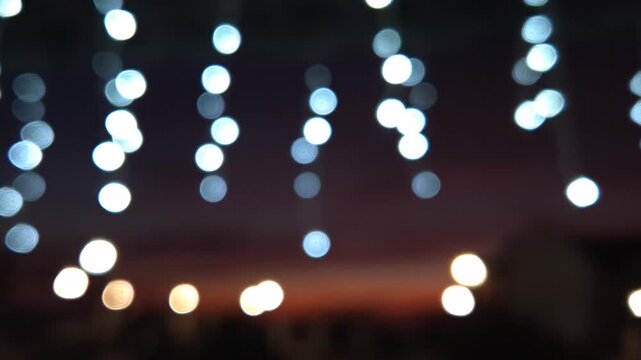 String lights bokeh, decorative glowing bulbs hanging at sunset against a dark evening sky. Handheld close-up shot.