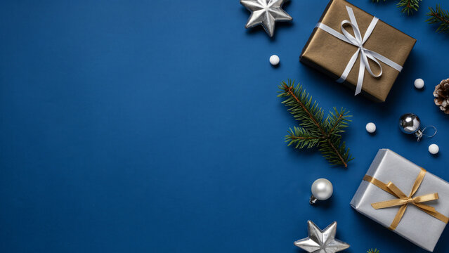 Elegant Christmas flatlay with gold and silver gift boxes, silver stars, fir branch, and baubles on classic blue background with copy space.