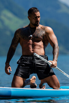 Shirtless tattooed man during summer paddleboarding. Tattooed SUP rider with muscular body. Tattooed male on lake with paddle. Full-length tattooed paddler against mountain scenery.