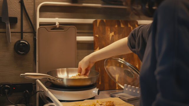Side view of woman placing raw seasoned chicken wings into hot frying pan on induction stove in modern loft interior. Food prep and cooking process on stainless steel countertop in warm home kitchen.