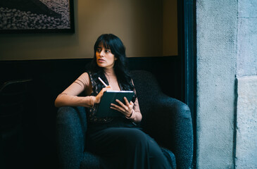 Thoughtful female student sitting in armchair holding digital tablet and stylus pen, contemplating online education course assignments e-learning research and study goals in city cafe © BullRun