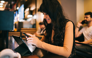 Smiling Caucasian female freelancer using smartphone next to digital tablet on desk, managing remote project task deadlines client communications and digital nomad workflow in cafe © BullRun