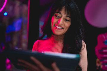 Close up portrait of smiling Caucasian female student looking down at digital tablet screen under neon lighting, browsing e-commerce retail store discount codes and mobile shopping app © BullRun