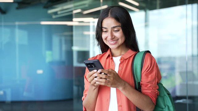 Happy young female student standing in modern university building smiling after reading good news on smartphone. Cheerful woman with backpack looking at phone, feeling pleased and joyful on campus.