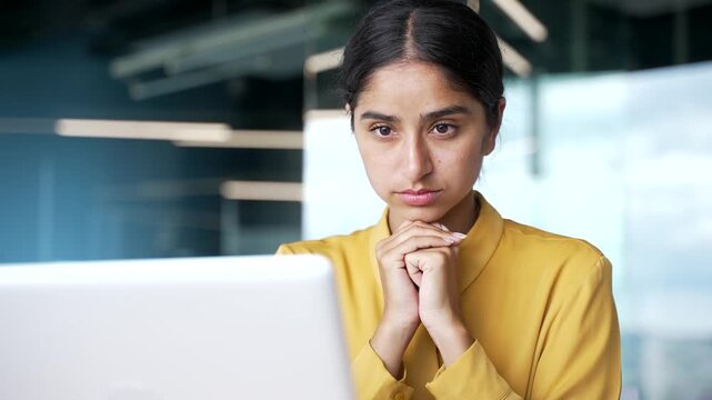 Close-up of young businesswoman watching video conference on laptop in modern office. Focused female employee listening to remote online training or learning course, taking part in virtual meeting.