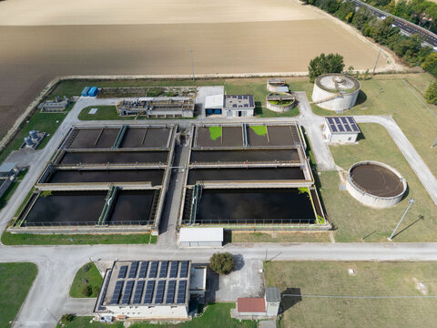 Aerial view of a wastewater treatment plant with rectangular sedimentation tanks, circular clarifiers, and solar panels on roofs, surrounded by agricultural fields in Marche, Italy.
