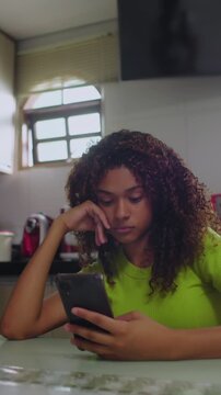 Hispanic teenage girl checking smartphone while sitting at kitchen table indoors