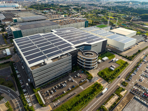 Aerial view of a large industrial warehouse facility with extensive rooftop solar panels and a distinctive spiral vehicle ramp in Zama, Kanagawa, Japan.