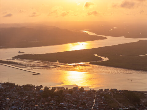 Aerial view of the river estuary at sunset with golden light reflecting on the water, surrounded by dense mangrove forests and the urban landscape of Cilacap, Central Java, Indonesia.