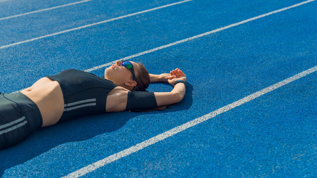 Woman athlete lying on running track relaxing after workout, female sprinter resting after training on sunny day, sports recovery, fitness, fatigue, rest and healthy active lifestyle concept