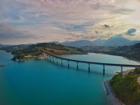 Aerial view of Lago di Cingoli with its long bridge crossing the turquoise water, surrounded by rolling hills and a cloudy sky in Cingoli, Marche, Italy.