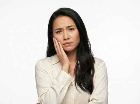 Asian woman touching her cheek with a concerned and worried expression on a white backdrop.