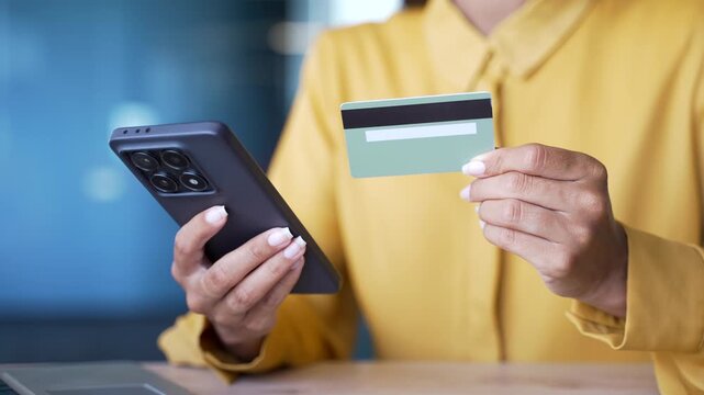 Close-up of female hands holding smartphone and credit card making online payment in office. Businesswoman entering data for mobile banking, digital transaction, or e-commerce purchase at workplace.