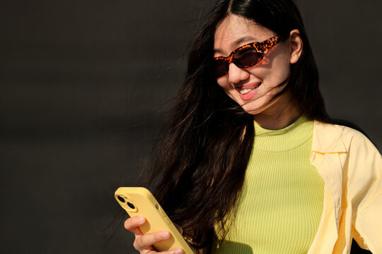 Portrait of young happy asian woman in sunglasses using mobile phone