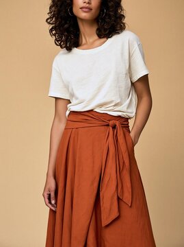 Young woman with curly hair wearing a white t-shirt and a long, flowing orange skirt. the skirt is tied at the waist with a knot, creating a flattering silhouette.