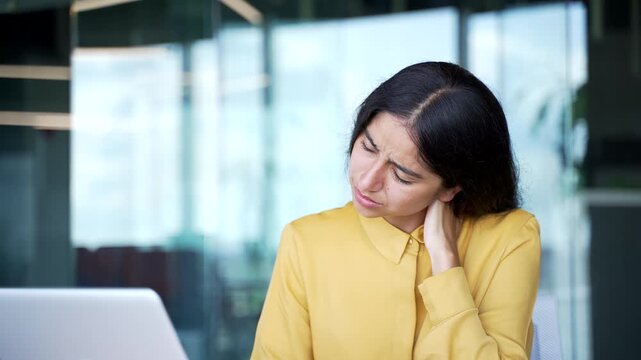 Close-up of young businesswoman massaging her painful neck while sitting in modern office. Tired female employee touching sore muscles, feeling stiffness and discomfort from tension and overwork.