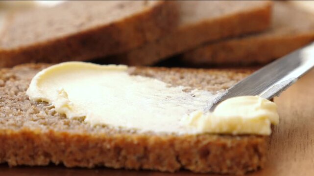 Butter spreads on whole wheat bread with metal knife. Close food preparation action moves from small dollop to smooth layer, creating fresh toast topping and simple breakfast comfort.
