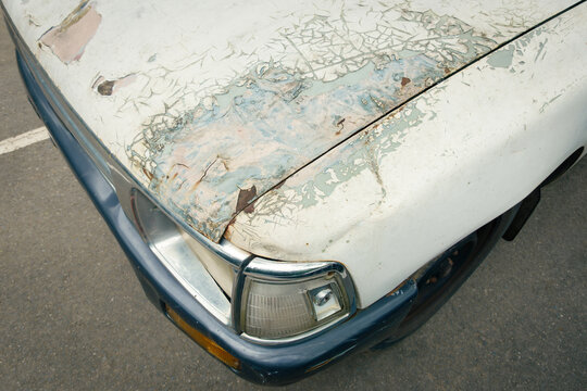 High-angle close-up shot of heavily cracked, peeling white paint and rusted metal on a vehicle hood, a cracked headlight assembly, grey bumper, and aged textures on an outdoor asphalt background