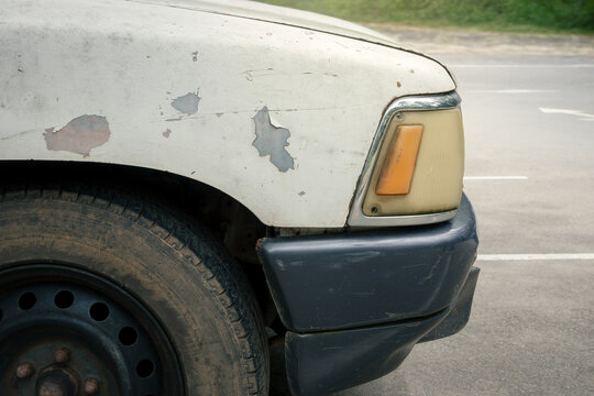 close-up shot of an old, white pickup truck with peeling body paint, a faded orange turn signal light, a scratched grey bumper, and a dirty black steel wheel parked on an asphalt road surface