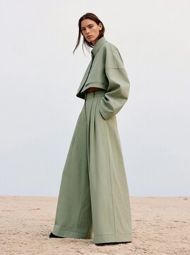 Young woman standing on a sandy beach. she is wearing a long, light green coat with a high collar and long sleeves. the coat is cinched at the waist with a belt, creating a flattering silhouette.