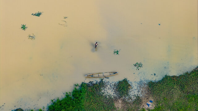 Aerial view of a lone fisherman standing in the shallow, murky waters of a river near a wooden boat, with lush green vegetation along the riverbank in Dambatta, Kano, Nigeria.