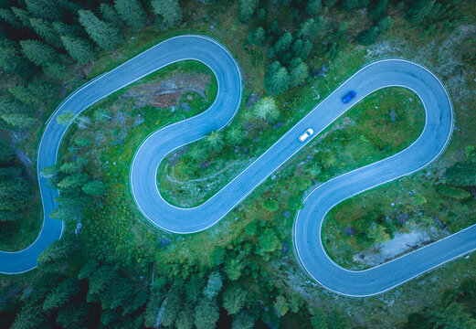 Aerial view of a dramatic mountain road with multiple tight hairpin bends winding through a dense alpine landscape with two vehicles driving along the asphalt. Italy.