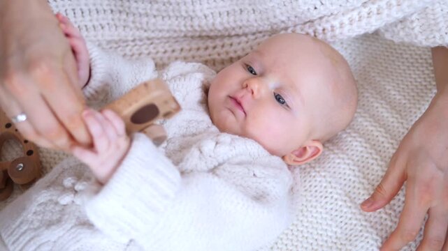 Infant holds wooden toy during gentle hand play. Baby lies on cozy knit blanket while adult hand steadies fingers, creating soft, peaceful moment of early sensory bonding and care.
