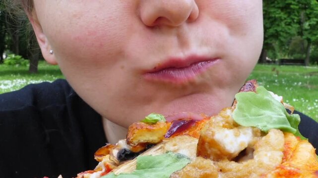 Woman munches pizza slice in green park setting, chewing steadily with simple pleasure. Tight framing emphasizes face, crust, and toppings while soft trees create natural backdrop.