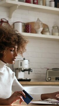 Young african american woman with afro hair sitting in her kitchen while shopping online, holding a credit card in one hand and typing on her laptop with the other to make a purchase