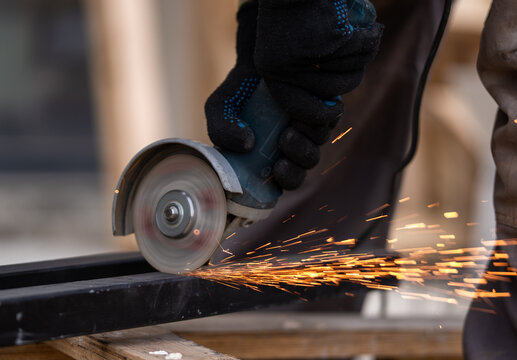  a worker in black protective gloves cutting a steel metal bar with an angle grinder. Stream of bright glowing sparks flying from the spinning cutting disc, industrial metalworking concept