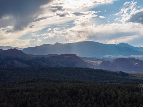 Aerial view of the majestic Rocky Mountains rising above a dense evergreen forest under a dramatic cloudy sky with soft sunlight filtering through Colorado, United States.