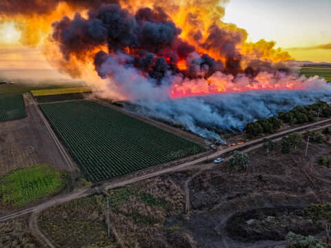 Aerial view of a sugarcane field fire with thick orange flames and billowing black smoke rising over agricultural land near a rural road in Home Hill, Queensland, Australia.