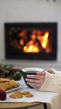 Hands Holding Warm Steaming Mug by Cozy Fireplace with Fresh Cookies, vertical video. Winter Hygge, Cozy Holiday Evening. Christmas Atmosphere