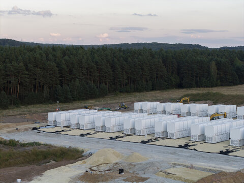 Aerial view of a battery energy storage system facility under construction with rows of white storage units, heavy machinery, and a forest backdrop in Trzebinia, Lesser Poland Voivode, Poland.