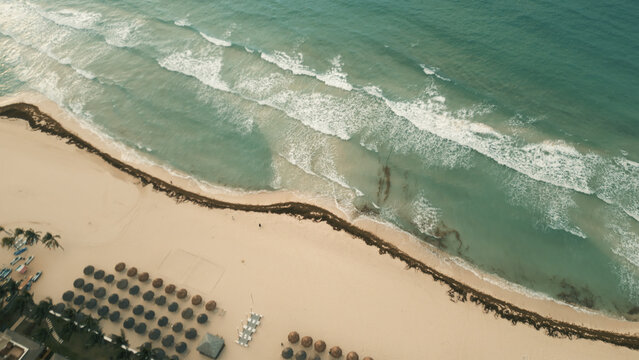 Aerial view of the beach with rows of straw umbrellas, turquoise waves, and seaweed on the sand in Playa del Carmen, Quintana Roo, Mexico.