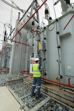 Conducting a substation inspection of the power transformer involves a technician in a hard hat holding a clipboard near wiring.