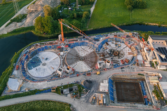 Aerial view of a construction site featuring circular concrete foundations, tower cranes, and excavation pits surrounded by green fields and a river.