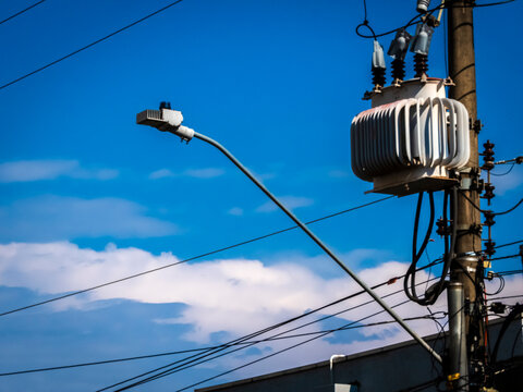 Electrical distribution transformer mounted on a concrete utility pole with a street light against a blue sky with soft white clouds, showing urban infrastructure and power grid in Brazil.