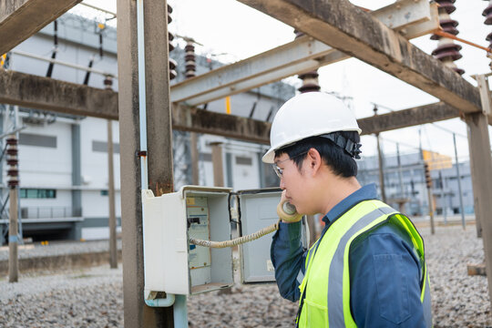 Electrical technician talks on a field telephone mounted in a weatherproof enclosure at an outdoor high voltage switchyard.