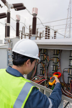 Checking a substation control cabinet during inspection, a technician measures current in wiring with a clamp meter beside a transmission tower.